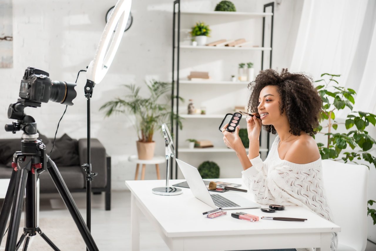 happy african american influencer in braces holding palette with eye shadow near digital camera